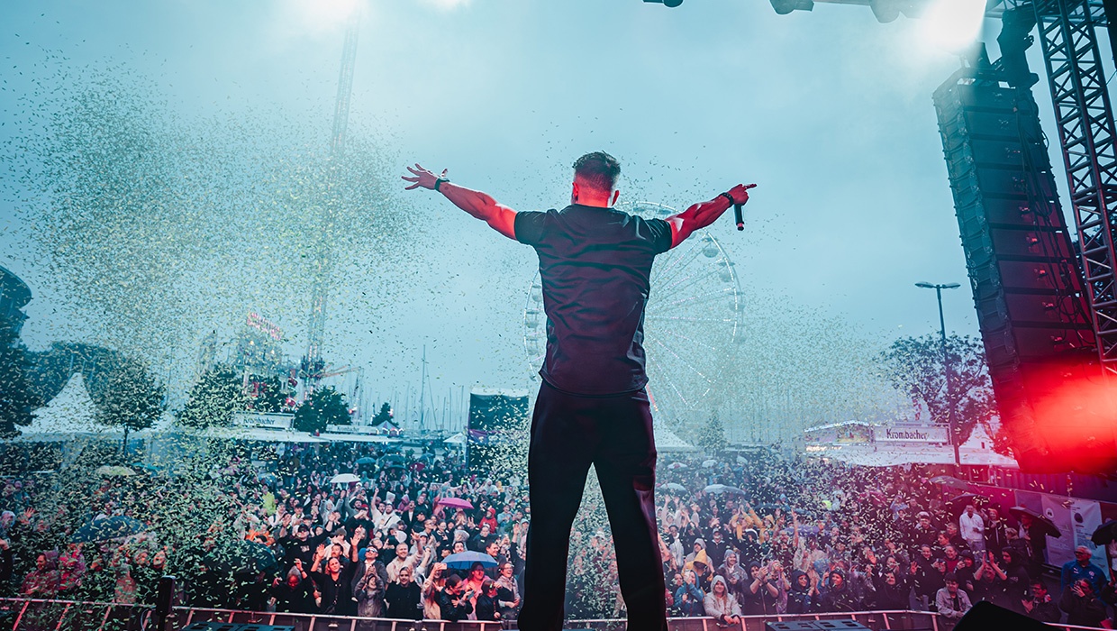Tiscore on stage with arms wide open, facing a massive festival crowd during a live performance.