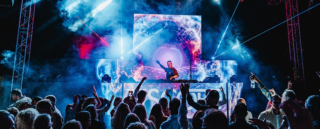 Tiscore performing a high-energy DJ set on a festival mainstage with lasers, smoke and a cheering crowd.